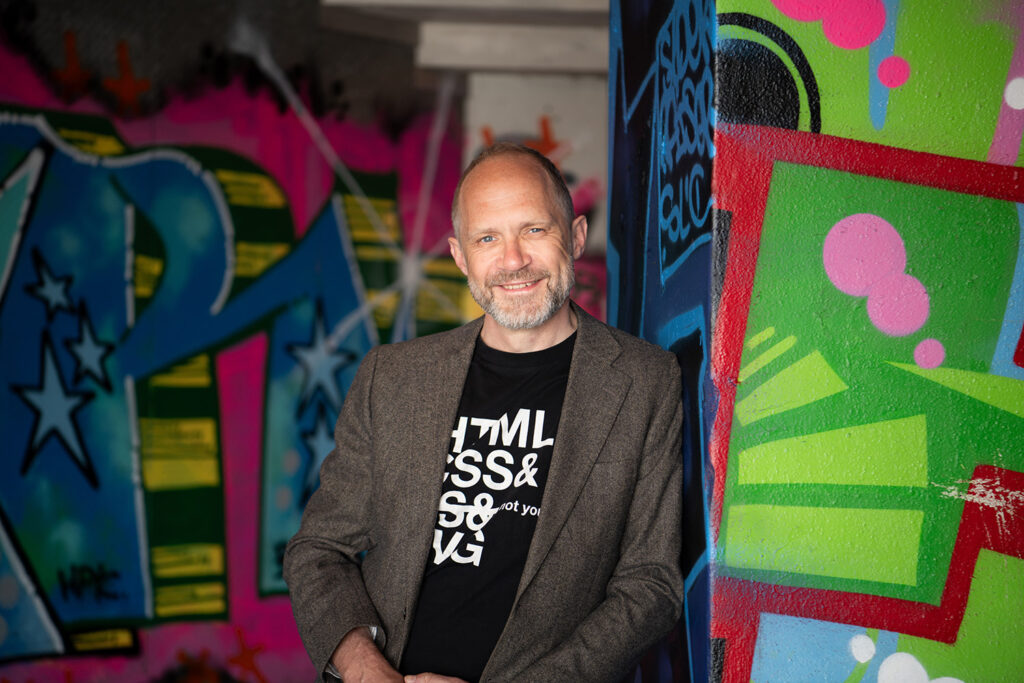Een zeldzame foto waarop Paul tandenbloot lacht. Portret van dichterbij. Paul glimlacht. Jasje en een t-shirt met daarop zichtbaar tekst: "HTML, CSS, JS, SVG" waarbij "JS" is doorgestreept. Op de muren kleurrijke graffiti.