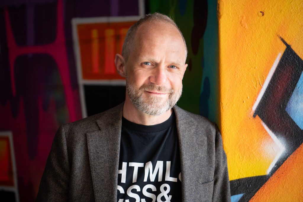 Portret van dichterbij. Paul glimlacht. Jasje en een t-shirt met daarop zichtbaar tekst: "HTML, CSS &"